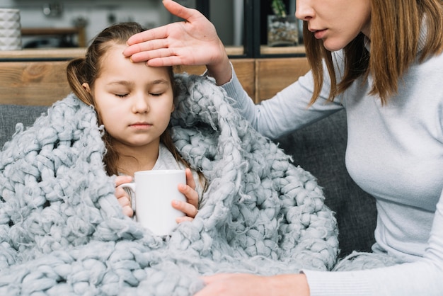 Close-up of a mother touching the forehead of her daughter holding white mug