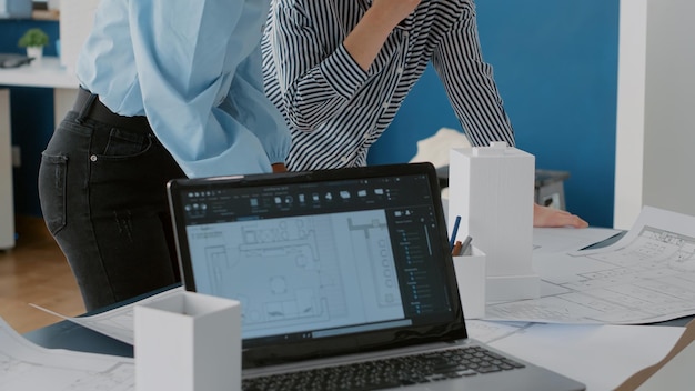 Close up of multi ethnic women working together to design construction blueprints for building model. Colleagues analyzing layout print plan, designing urban architectural project.