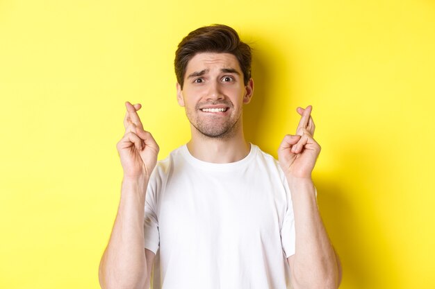 Close-up of nervous man making wish, holding fingers crossed and biting lip worried, standing over yellow background.