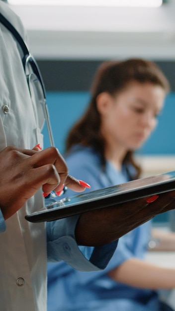 캐비닛의 의료 정보를 위해 터치 스크린이 있는 태블릿을 사용하는 아프리카계 미국인 의사의 클로즈업. 의료를 위한 현대적인 장치를 들고 흰색 코트와 청진기를 가진 흑인 위생병 무료 사진