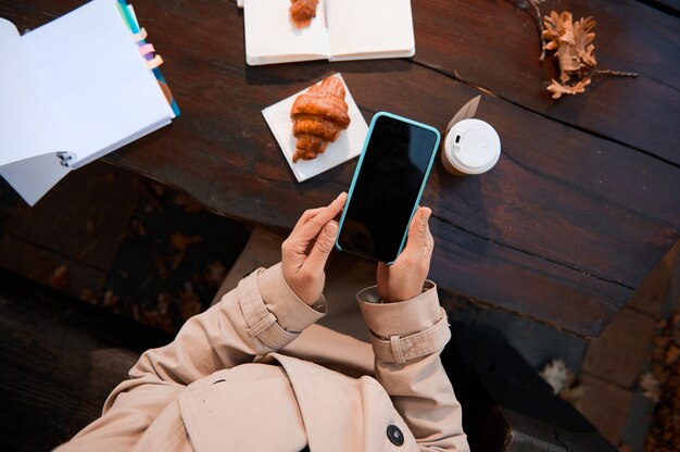 開いた​メモ帳​と​コピー​ブック​と​おいしい​朝食​と​木製​の​テーブル​の​背景​に​コピー​スペース​の​ため​の​空白​の​黒い​画面​と​携帯​電話​を​使用して​認識​できない​人​の​手​の​クローズアップ