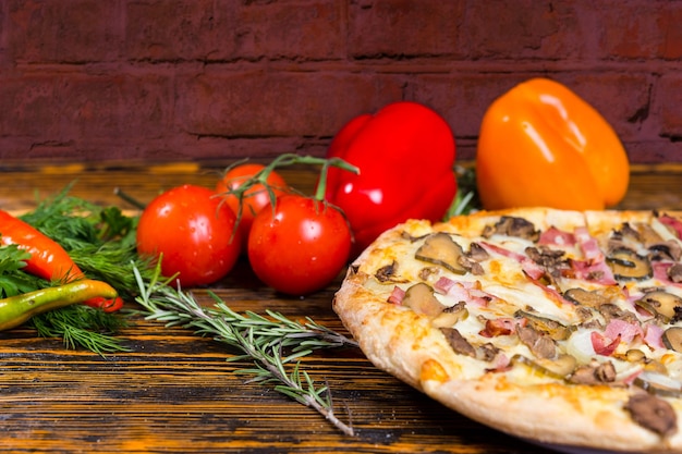 ハム、マッシュルーム、タマネギ、ピクルスが近くにあるおいしいピザのクローズアップは、木製のテーブルにコショウ、トマト、ローズマリーの小枝やその他の野菜です Premium写真