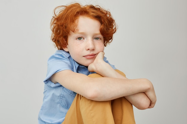 Бесплатное фото Крупным планом забавный маленький мальчик с красными волнистыми волосами, сидя на полу, держа лицо рукой
