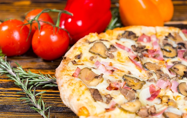 근처에 햄, 버섯, 양파, 피클이 있는 맛있는 피자를 가까이서 나무 탁자에 있는 고추, 토마토, 기타 야채