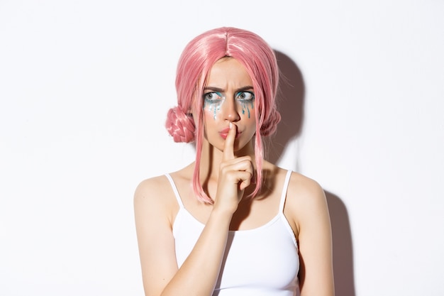 Бесплатное фото Крупный план взволнованной привлекательной девушки в розовых аниме-волосах, показывающей знак «шуш», прижатой указательным пальцем к губам и озабоченной. женщина поделится секретом, стоя.