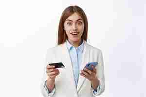 Бесплатное фото Крупный план молодой деловой женщины в костюме, легко оплачиваемой онлайн с пластиковой кредитной картой и смартфоном, делающей покупки в интернете, держащей мобильный телефон, удивленной спереди, белая стена