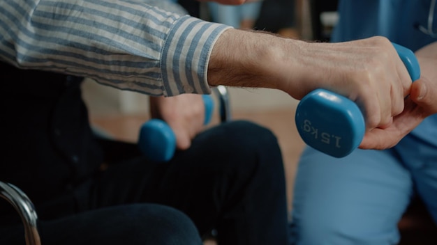 Close up of old patient with disability holding dumbbells and doing physical exercises with help from nurse in nursing home. Healthcare specialist supporting man in wheelchair with recovery