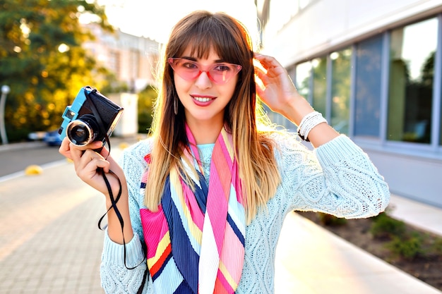 Close up outdoor city portrait of magnificent young pretty woman holding retro vintage film camera, wearing pastel sweater sunglasses and scarf, evening sunlight.