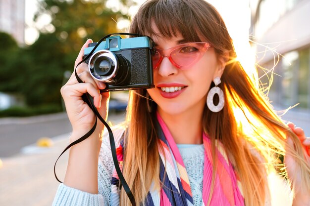 Close up outdoor city portrait of magnificent young pretty woman holding retro vintage film camera, wearing pastel sweater sunglasses and scarf, evening sunlight.