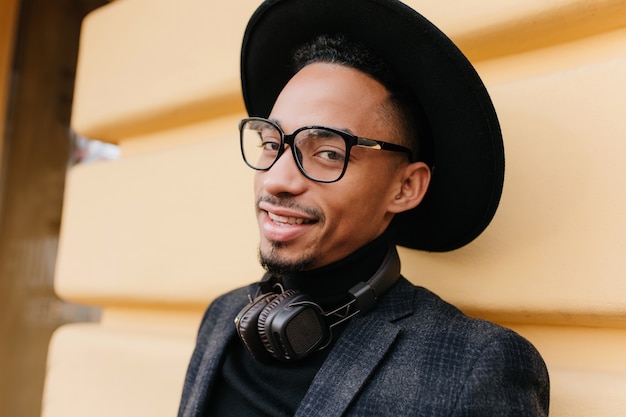 Close-up outdoor photo of carefree man in stylish dark clothes posing near old building. Handsome african guy standing in front of yellow wall.