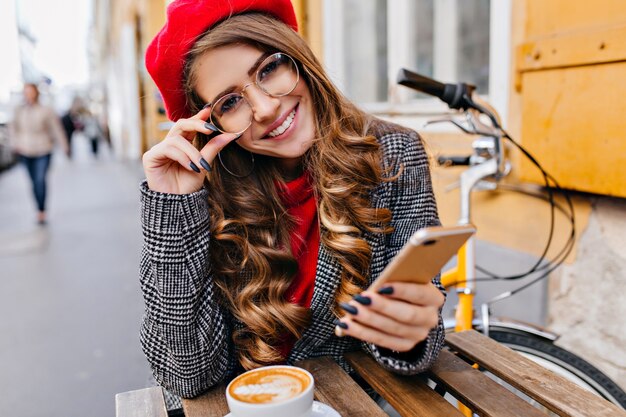 Close-up outdoor photo of lovely female model in glasses drinking hot cappuccino on urban background