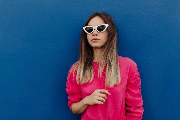 Close up outdoor portrait of charming young woman wearing pink shirt and white sunglasses posing over isolated blue wall