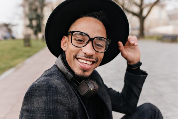 Close-up outdoor portrait of excited young man with brown skin wears glasses. African bearded boy in hat expressing positive emotions.