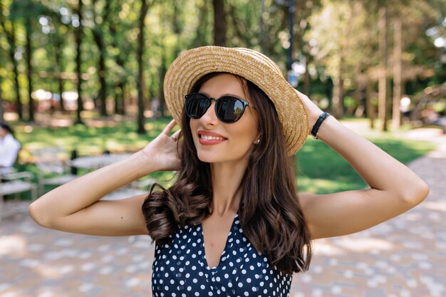 Close up outdoor portrait of lovely friendly stylish woman wearing sunglasses and hat in summer dress is holding hat