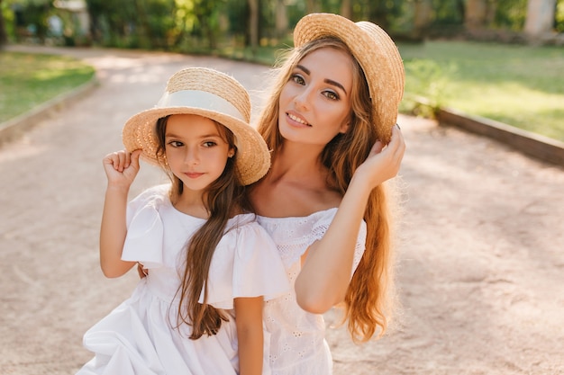 Close-up outdoor portrait of pretty dark-eyed girl looking away while posing with mom in the park. Charming long-haired woman with trendy straw hat playing with hair, standing near daughter on road.