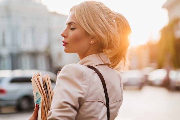 Close-up outdoor portrait of romantic fair-haired girl holding newspaper and posing with eyes closed early in evening