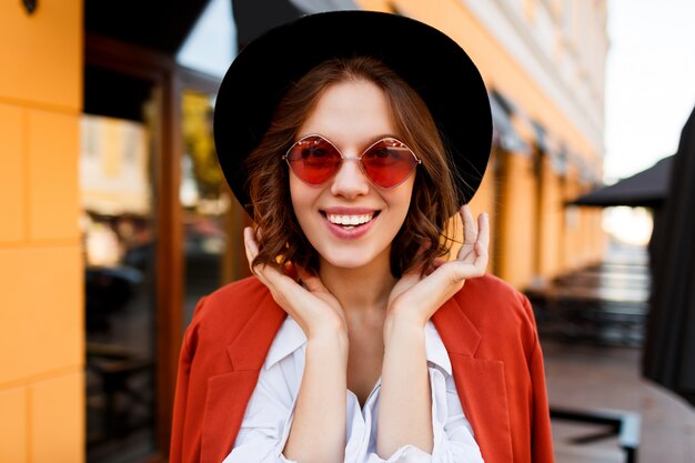 かわいいオレンジ色のサングラス、ジャケット、黒い帽子で笑顔のヨーロッパの女の子の屋外のポートレートを閉じます。秋のファッション。