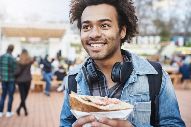 アフロの髪型と幸せな感情的な若い浅黒い男性、首とデニムのコートの上にヘッドフォンを着用、サンドイッチを押しながら市のお祭りにいる間よそ見の屋外のクローズアップショット