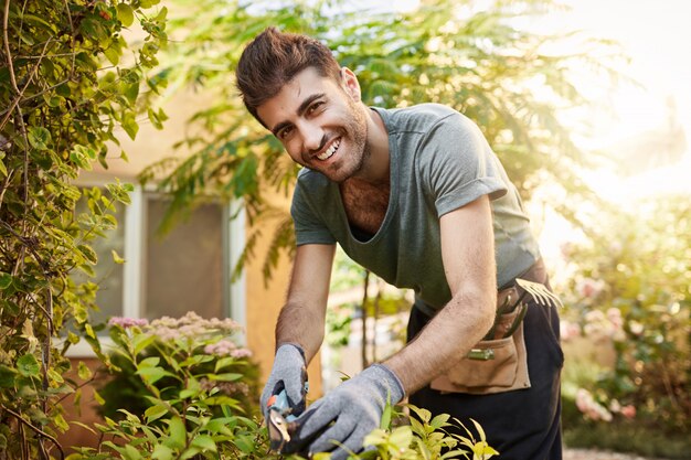 Close up outdoors portrait of beautiful cheerful bearded caucasian farmer in blue shirt and gloves smiling , working with garden tools on his farm near countryside house