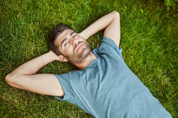 Close up outdoors portrait of young attractive mature bearded hispanic man in blue t shirt looking in camera, lying on ground with relaxed face expression.