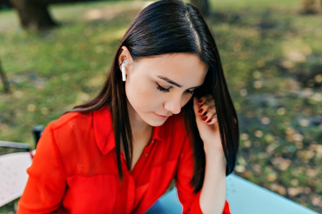 Close up outside photo of dark haired woman is wearing red blouse is listening music in open air terrace