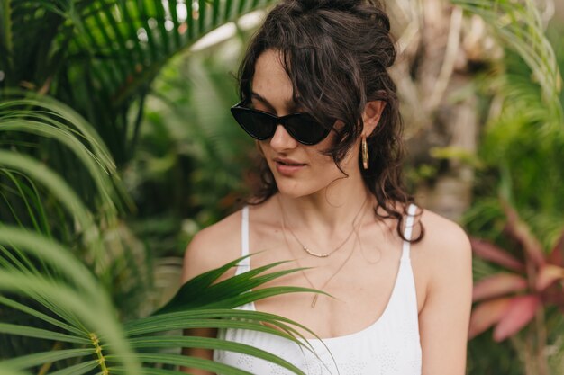 Close up outside portrait of adorable happy girl with blond curly hair wearing stylish modern yellow glasses  playing with hair and looking down on wall of green plants