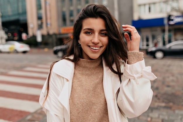 Close up outside portrait of smiling pretty woman with dark hair wearing beige sweater and white coat walking on the street