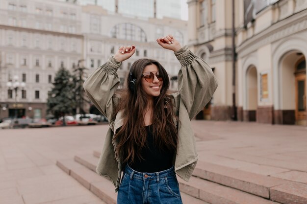 Close up outside portrait of smiling pretty woman with long hair wearing jacket walking in the city with headphones enjoying favorite music