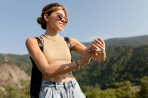 Close up outside portrait of stylish pretty girl in sunglasses is looking at smart watch and walking with backpack in sunny day on background of green hills