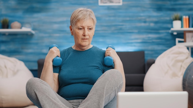 Free photo close up of pensioner training with dumbbells on yoga mat, looking at laptop with workout video. retired woman using weights lifting to exercise and following online workout on computer