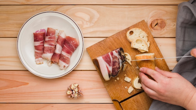 Primo piano di una persona che taglia la fetta di pane con pancetta arrotolata sul piatto bianco sopra la tavola di legno