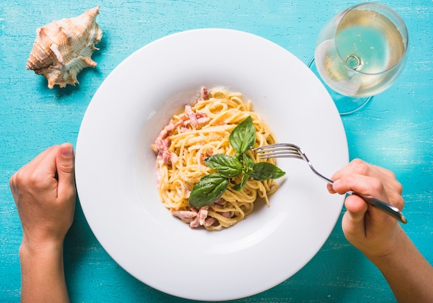 Close-up of a person eating spaghetti with wineglass and conch on turquoise backdrop