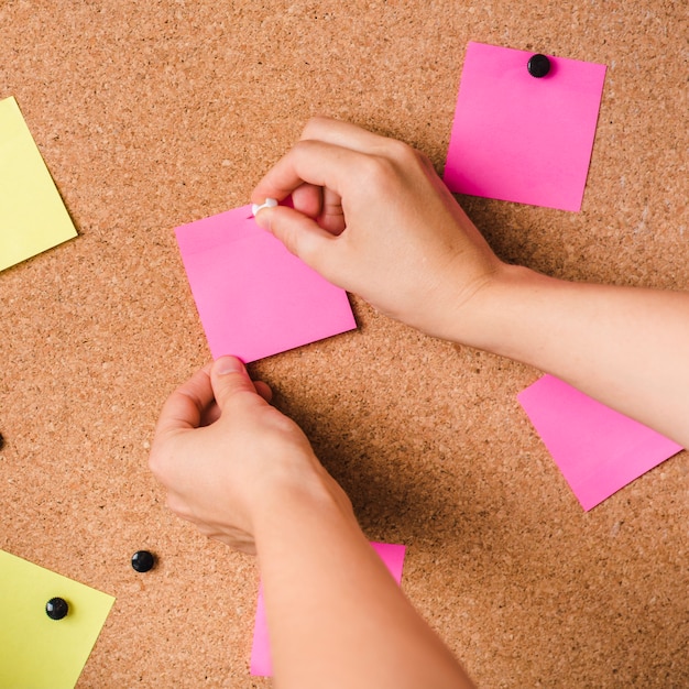 Close-up of a person fixing pink adhesive note with thumbtack on corkboard
