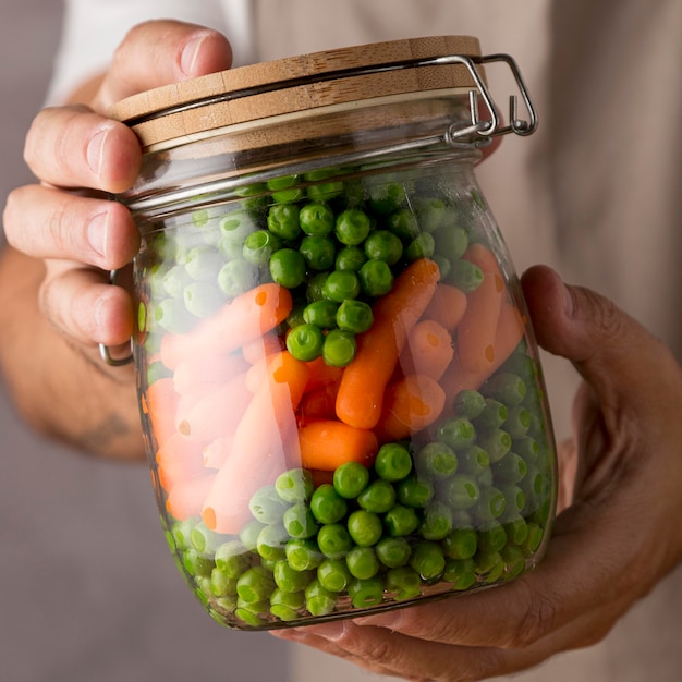 Free photo close-up of person holding peas and baby carrots in glass jar