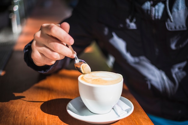 Close-up of a person holding spoon over the cappuccino or latte with frothy foam