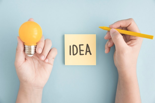 Close-up of a person holding yellow crayon and light bulb with idea text on sticky note over the grey background