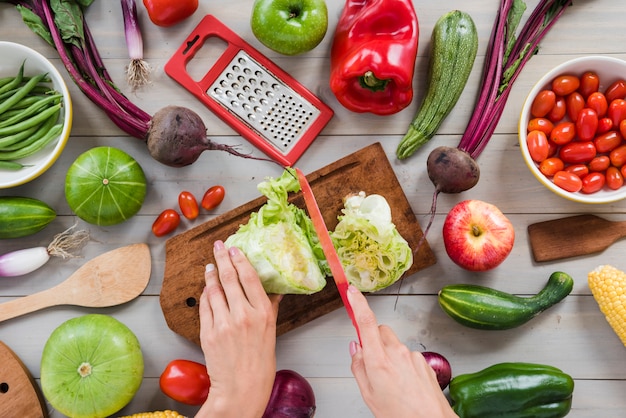 Close-up of a person's hand cutting cabbage with knife on chopping board surrounded with vegetables on table