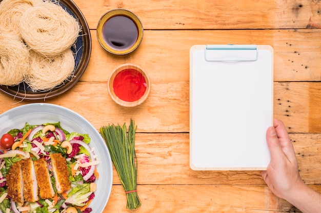 Close-up of a person's hand holding clipboard near the traditional thai food on wooden table