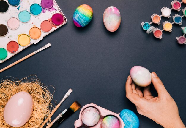 Close-up of a person's hand holding the painted egg with paint color and brushes on black background