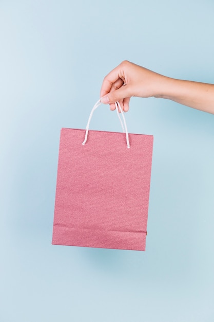 Close-up of a person's hand holding pink paper shopping bag on blue backdrop