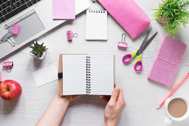 Close-up of a person's hand holding spiral notepad with office stationeries on wooden desk