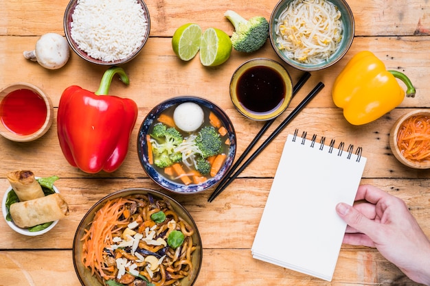 Close-up of a person's hand holding spiral notepad with thai food on table