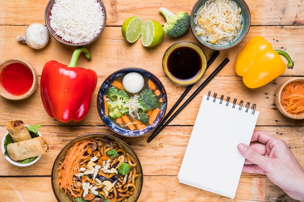 Close-up of a person's hand holding spiral notepad with thai food on table