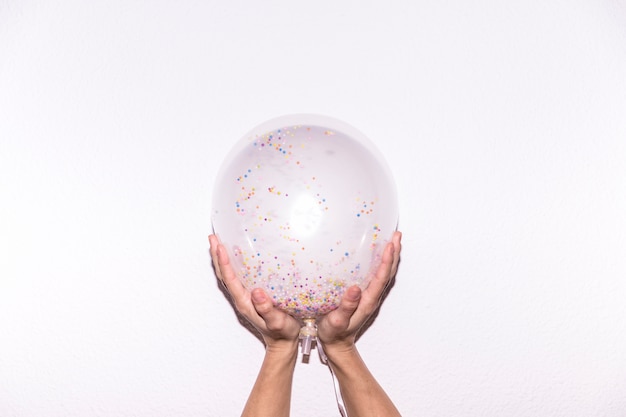 Close-up of a person's hand holding transparent white balloon with colorful sprinkles against white backdrop