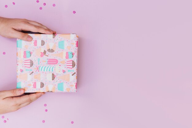 Close-up of a person's hand holding wrapped gift box with confetti on pink backdrop