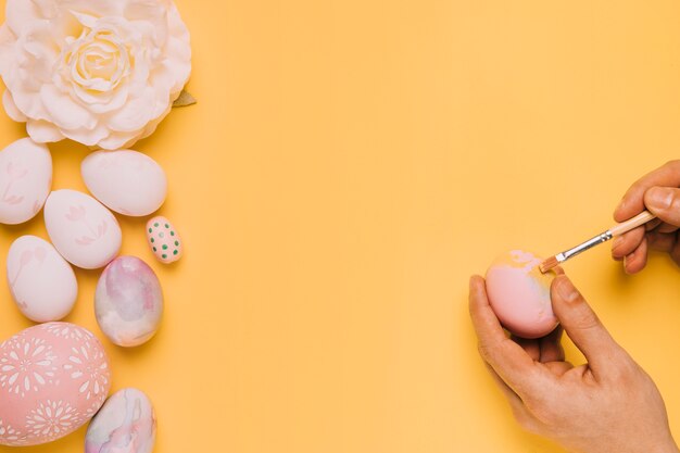 Close-up of a person's hand painting the egg with paint brush on yellow backdrop