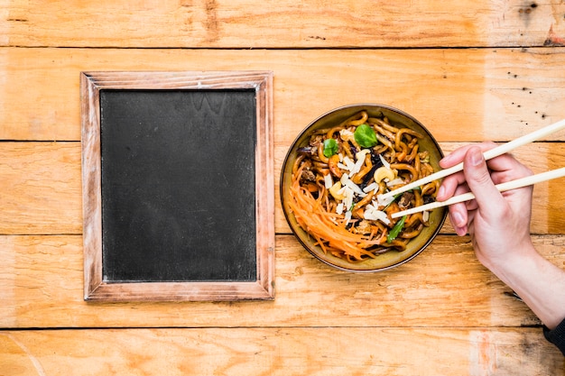 Close-up of a person's hand picking the noodles with chopsticks near the blank slate on table