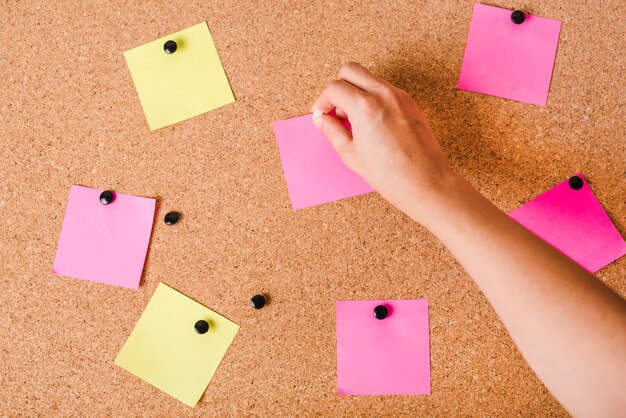 Close-up of a person's hand putting blank notepad on cork board with push pin