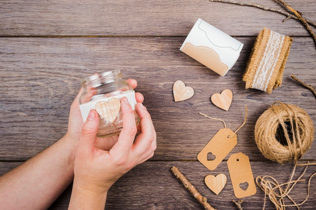 Close-up of a person's hand sticking the wooden heart on transparent bottle over the wooden desk