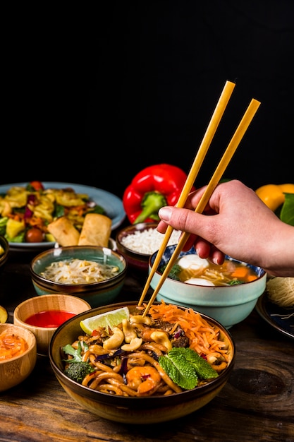 Close-up of a person's hand taking thai food with chopsticks against black background